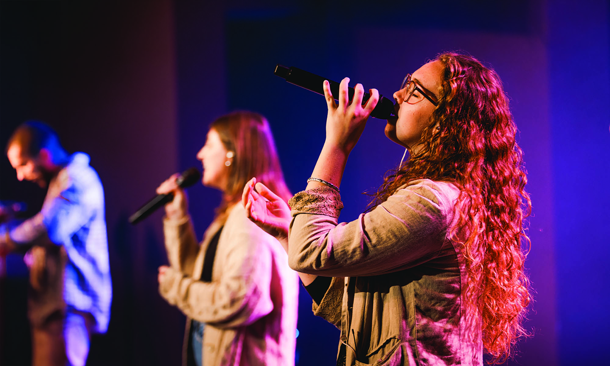 Anderson University students leading worship, singing into microphones with hands raised on a stage lit in blue and magenta.