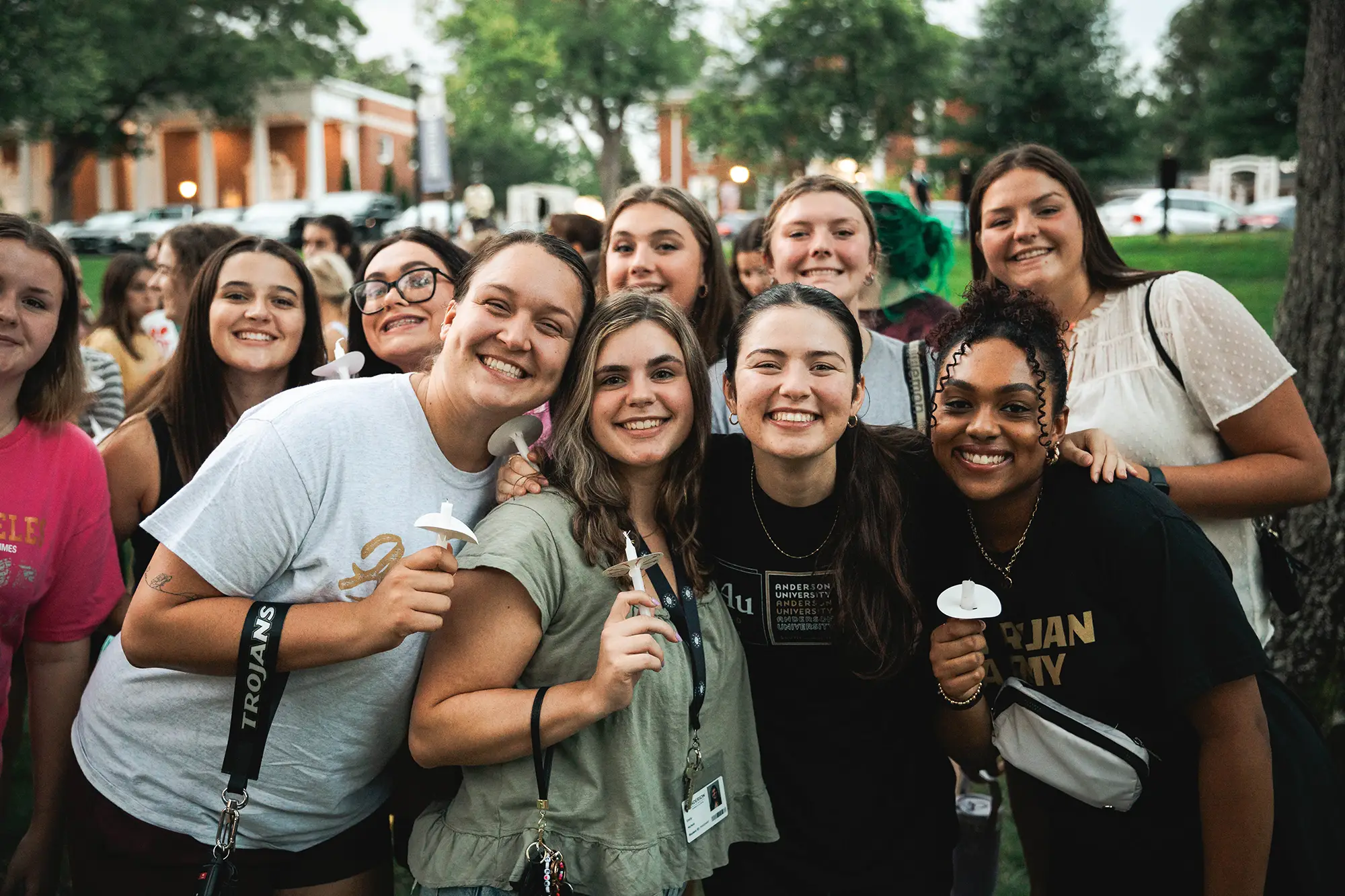 A joyful group of Anderson University students gathered together outdoors on campus at dusk holding candles.