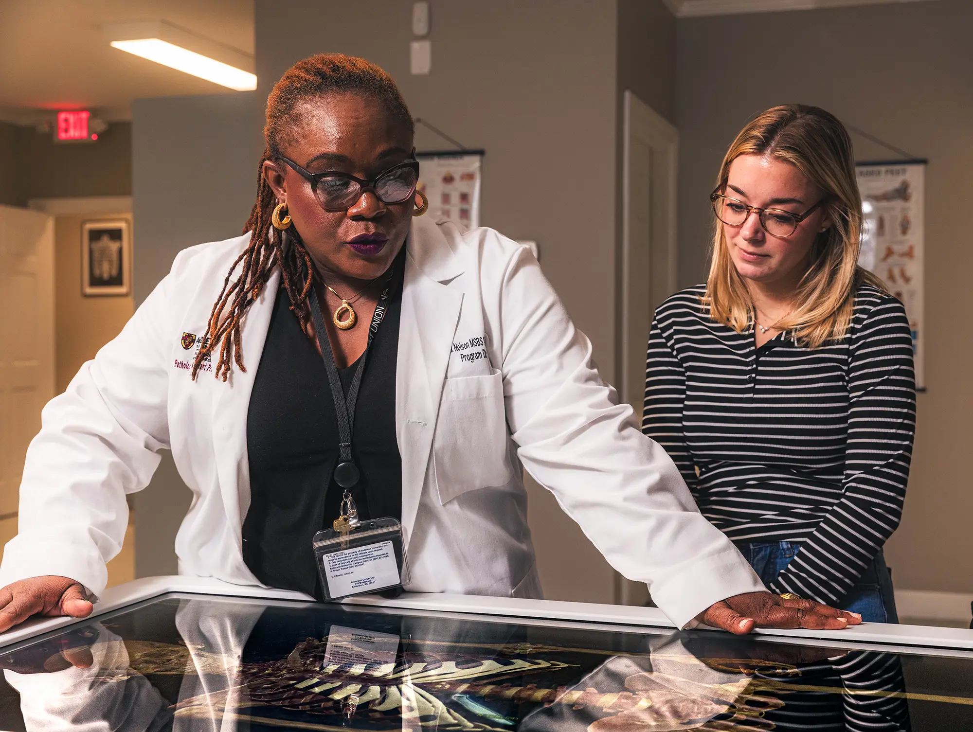 An Anderson University professor in a white lab coat mentoring a student over an illuminated anatomical display.